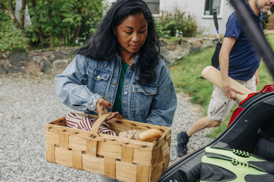 Woman wearing denim jacket loading food basket in electric car trunk