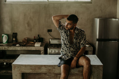 Pensive young ethnic male in casual clothes looking away and sitting on counter in kitchen