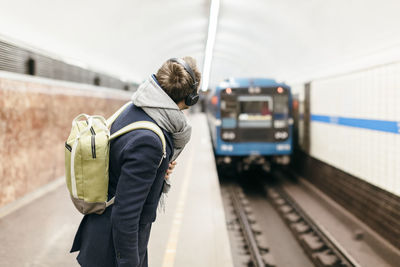 Handsome man in coat with green backpack and headphones looks after the departing metro train. 