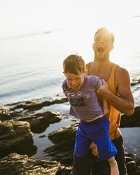 Happy father carrying son while standing on rocks at beach against sky during sunset