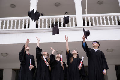 Students in university gowns throwing mortarboards while standing against building