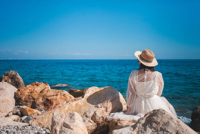 Rear view of woman sitting on rock by sea against clear blue sky