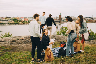 Happy friends with bags talking on field by lake against sky