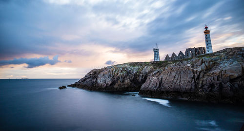 Lighthouse on sea by buildings against sky during sunset