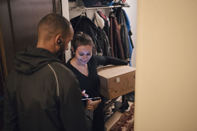 Smiling woman signing on mobile phone for delivery from messenger at doorway