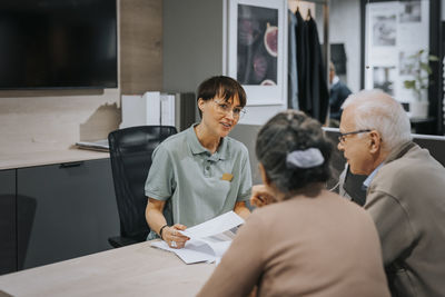 Smiling female interior designer explaining senior couple over brochure at desk in store