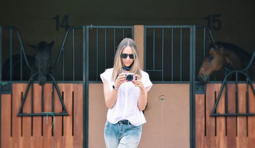 Young woman holding camera with horses in background at stable