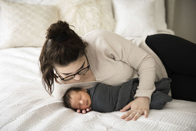 Hipster millennial mom snuggles swaddled newborn on white bed