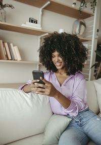 Smiling afro woman using smart phone sitting on sofa at home