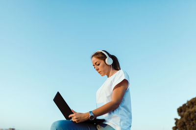 Low angle view of young woman using laptop against clear sky