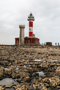 Lighthouse on beach by buildings against sky