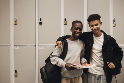 Portrait of smiling teenage boys wearing backpacks while standing against locker in school