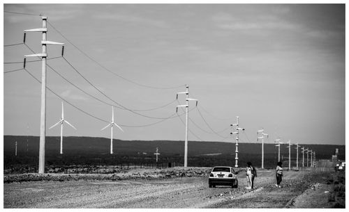 Car on dirt road by electricity pylons and windmills against sky during sunny day