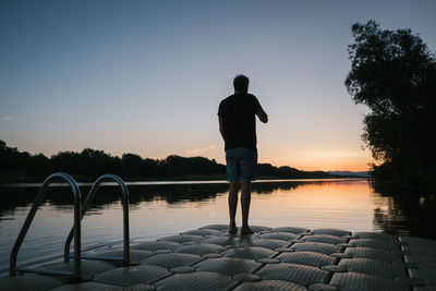 Rear view of silhouette man standing on pier over lake against sky during sunset