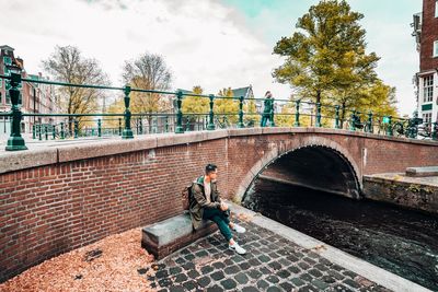 Man on retaining wall by bridge against sky