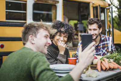 Smiling man showing smart phone to friends sitting at table against caravan