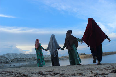Rear view of women holding hands while standing at beach against sky