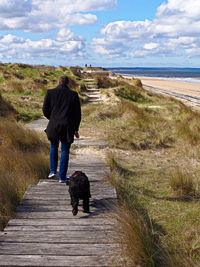 Rear view of man with dog walking on walkway amidst grass at beach against sky