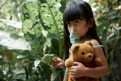 Girl breathing with nebulizer while standing with teddy bear by plants