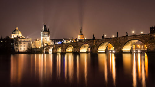 Illuminated bridge over river against buildings at night