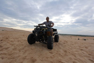 Man riding quadbike at desert against cloudy sky