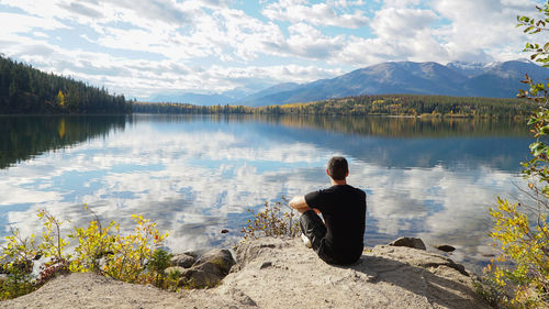 Rear view of man sitting by lake against sky