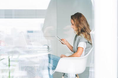 Young adult woman with long hair working on laptop using smartphone sitting on kitchen at the home