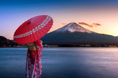 Woman wearing traditional clothing holding umbrella while standing by lake during sunset