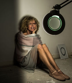 Smiling mature woman wrapped in a blanket sitting on floor while looking at lens against wall