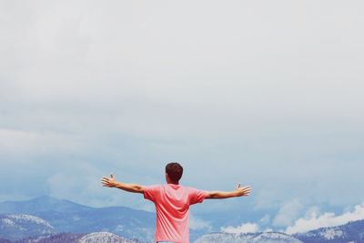 Rear view of man with arms outstretched standing at mountains against cloudy sky