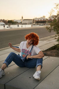Full length of young woman sitting in city against sky during sunset