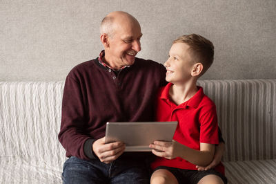 A happy grandfather hugs his grandson. they hold a tablet in their hands and look at each other
