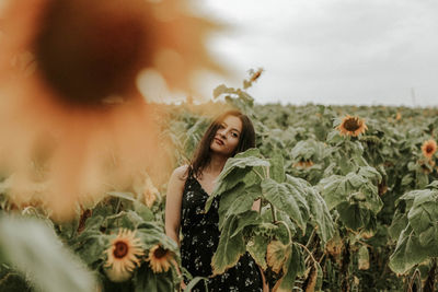 Young woman standing amidst sunflowers on field against sky