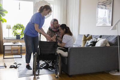 Mother and female caregiver assisting daughter while sitting on sofa in living room at home