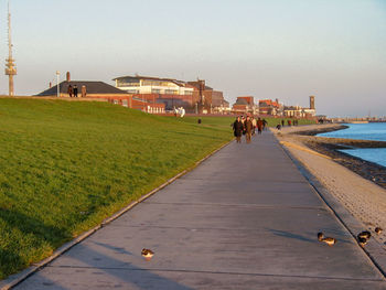 People walking on shore by city against clear sky