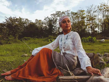 Thoughtful young woman wearing traditional clothing sitting on land against sky