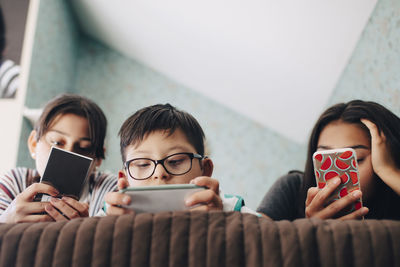 Low angle view of boy looking at digital tablet while lying with sisters using mobile phone on bed at home