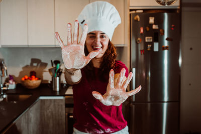 Portrait of woman wearing chef hat showing messy hands while standing in kitchen