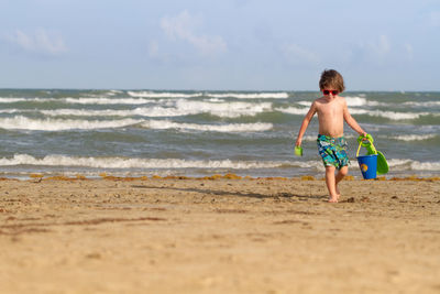 Shirtless boy holding toys while walking at beach