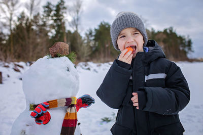 Boy in hat bites carrot, which was the nose of snowman, smiling. winter family weekend, lifestyle