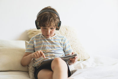 Cute boy lying on the bed at home and playing with pc tablet with wireless headphones