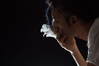 Close-up of man with hand covering mouth emitting smoke against black background