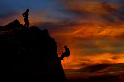 Silhouette people on rock climging against dramatic sky during sunset