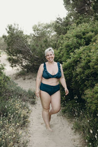 Positive curvy female with short hair in swimwear walking looking at camera on sandy path on beach and enjoying summer