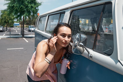 Young woman with water bottle adjusts make up looking in the mirror of a camper van in urban parking