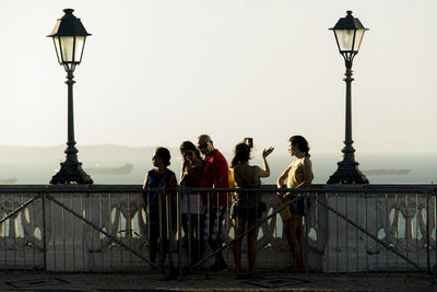 People standing on street light by bridge against sky