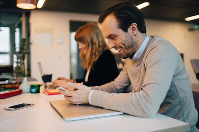 Side view of smiling businessman using smart phone while sitting by businesswoman at desk in creative office