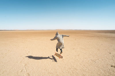 Rear view of man skateboarding on sand against clear blue sky
