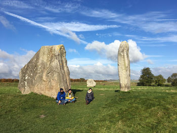 Friends sitting on grassy field against sky at avebury stone circle