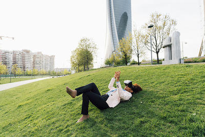 Businesswoman wearing virtual reality simulator lying on grass gesturing in city